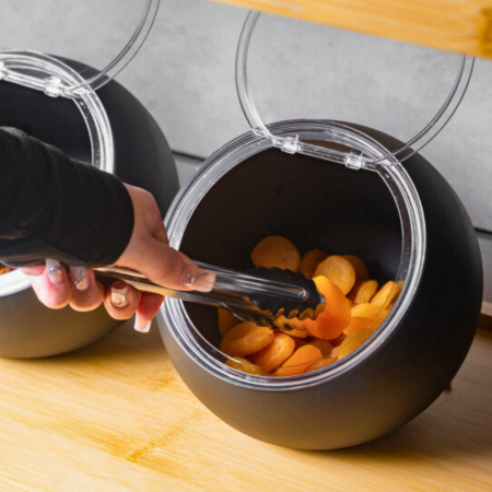 Person using tongs to take dried apricots from a black container on a wooden surface.