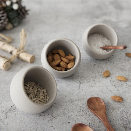Three small ceramic bowls filled with different types of seeds and a wooden spoon on a gray surface.
