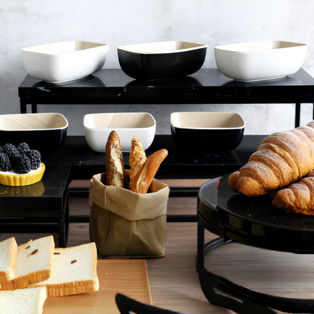 Ceramic bowls on a black riser with bread and pastries on a wooden surface.